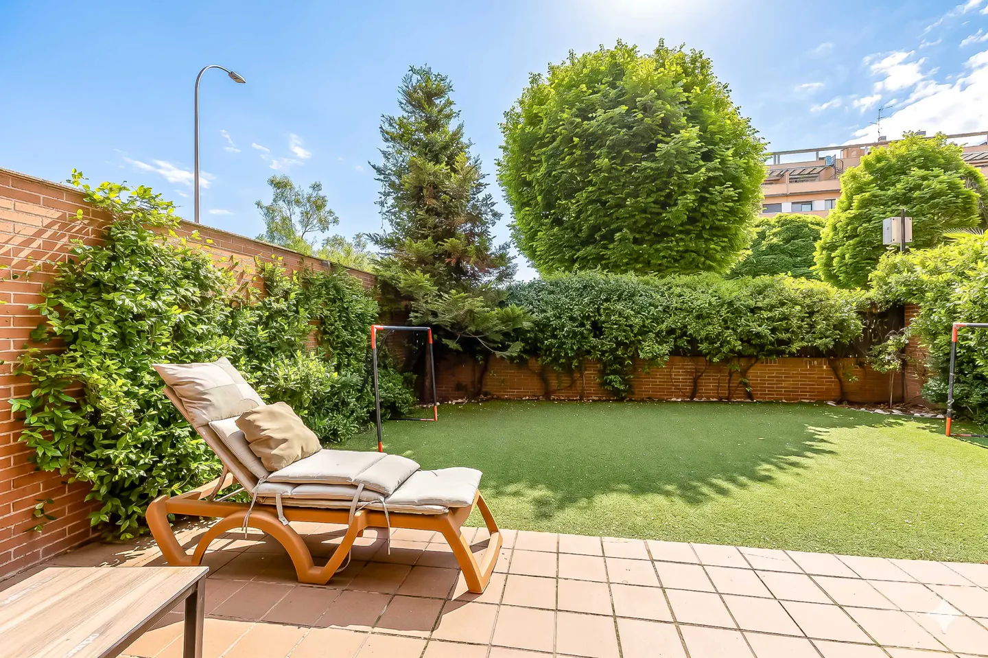 A backyard with a lounge chair, green grass, trees, and a brick wall on a sunny day.