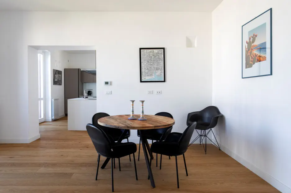 Bright dining area with wood floors, white walls, and a round wood table with black chairs. Artwork hangs on the wall. Kitchen visible in background.