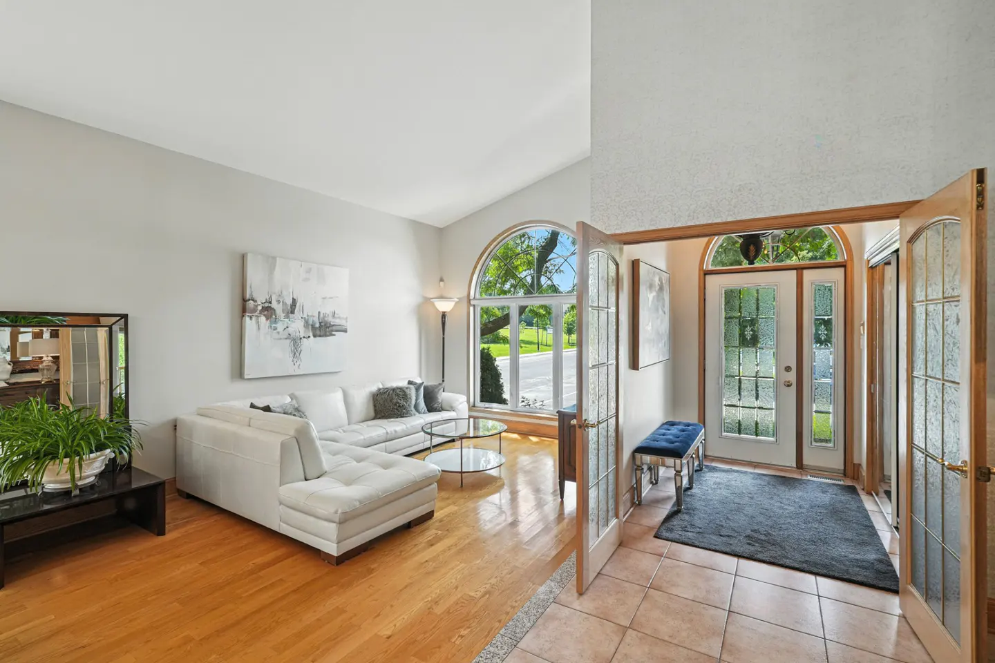 Bright foyer with hardwood floors, white walls, and a white sectional sofa. Double doors lead to the outside. A blue bench sits near the entrance.