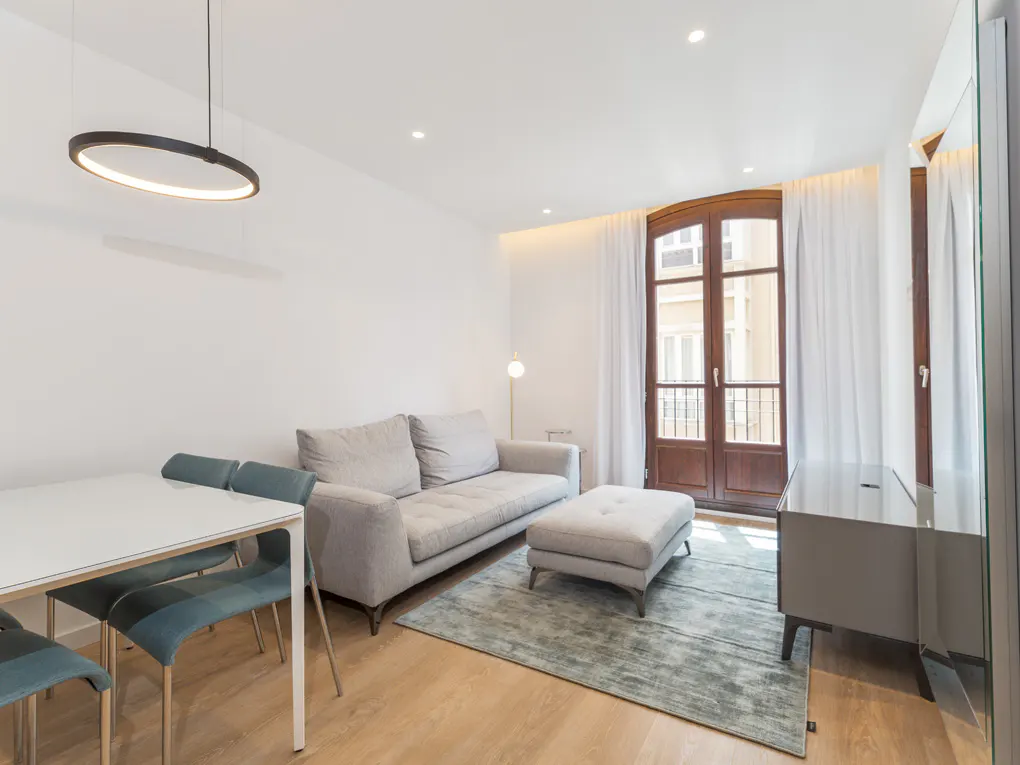 Bright living room with white walls, wood floors, and a gray sofa. A white table with blue chairs sits near a modern, circular light fixture. A large window with white curtains provides natural light.