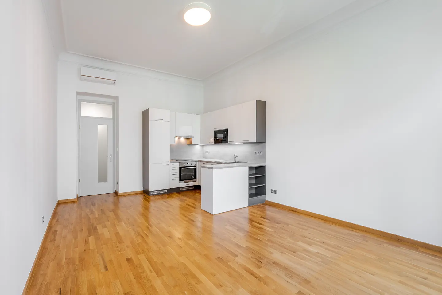 Bright, empty apartment with hardwood floors, white walls, and a modern white and gray kitchen. A door is visible on the left.
