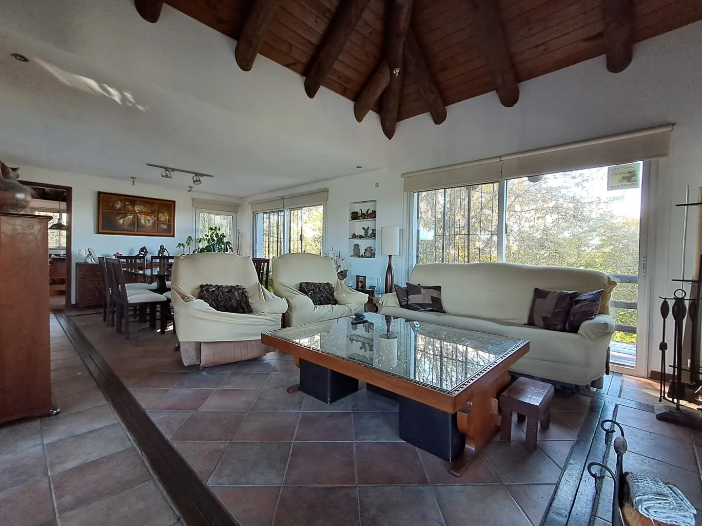 Living room with cream sofa and chairs, glass coffee table, and terracotta tile floor. Large windows overlook trees. Wooden ceiling.