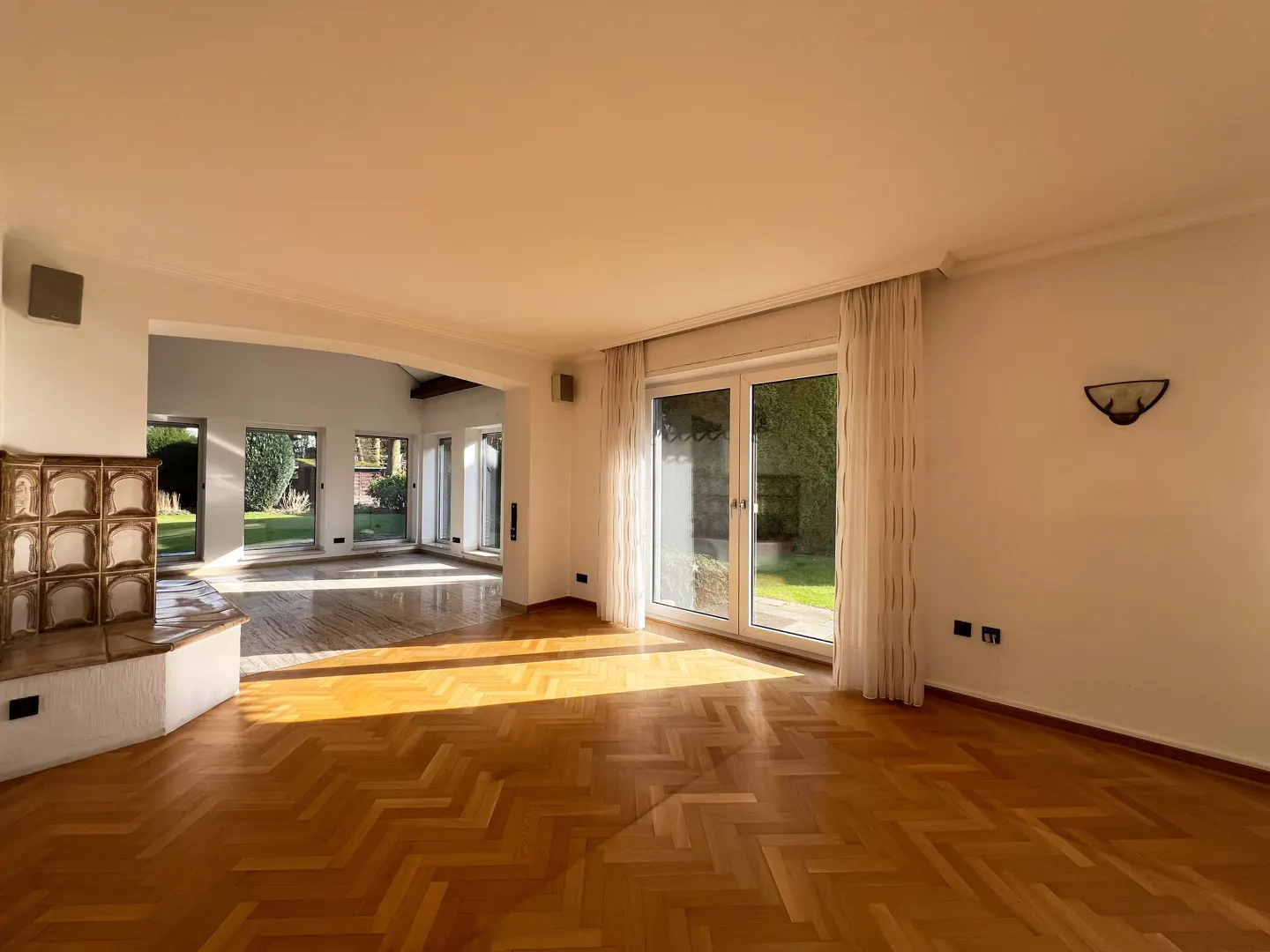 Bright, empty living room with herringbone wood floors, white walls, and a tiled fireplace. Sunlight streams through glass doors to a green garden.