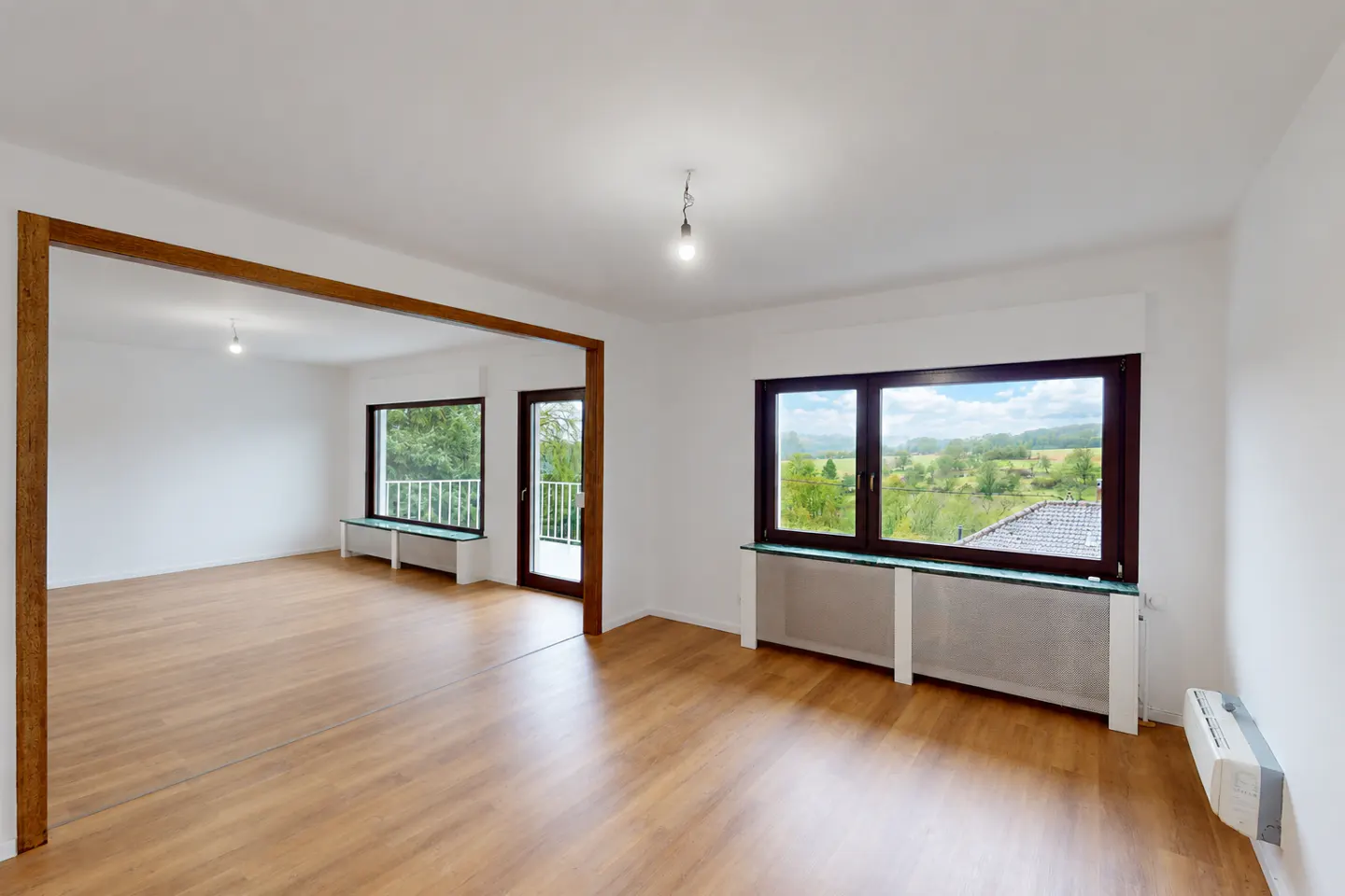 Bright, empty room with wood floors and white walls. Large windows offer views of greenery. A wooden frame separates the space.
