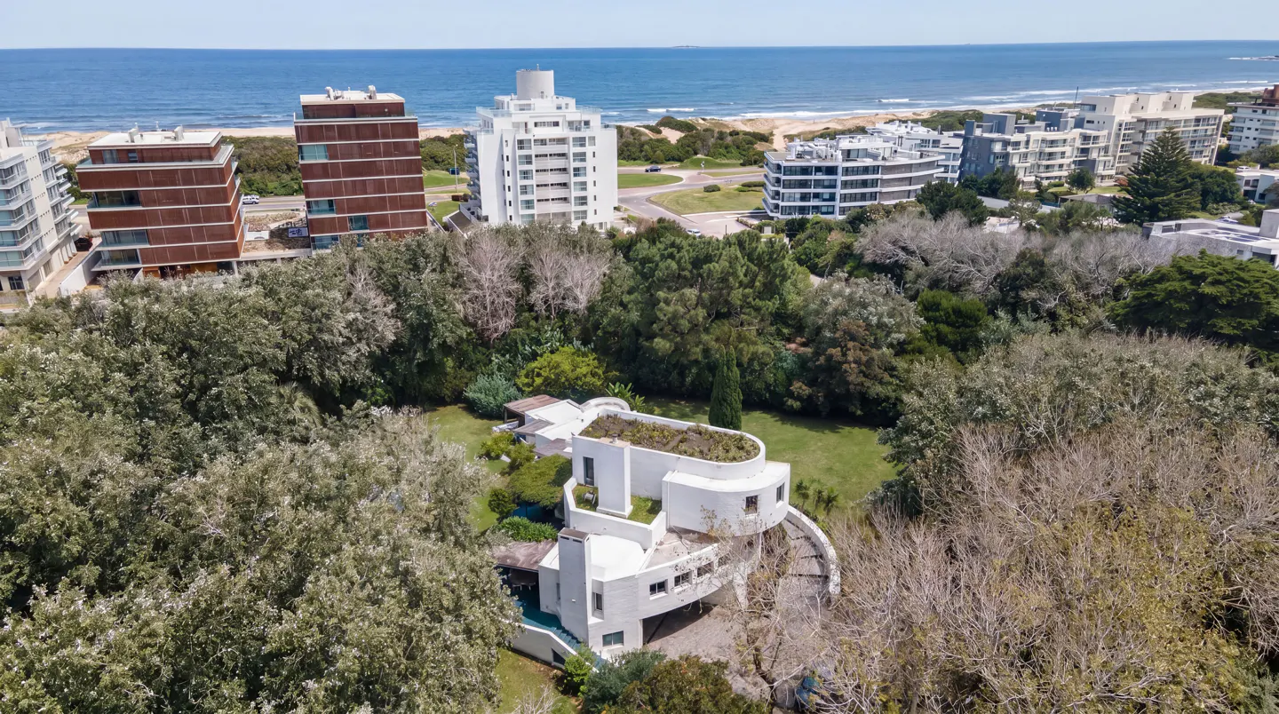 Aerial view of a modern white house with a green roof, surrounded by trees, with the ocean and buildings in the background.