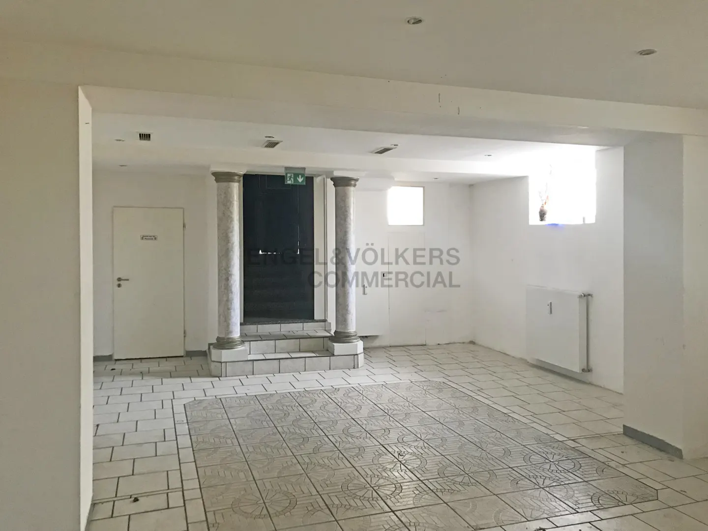 A bright, empty commercial space with white walls, tiled floors, and two marble-like columns leading to a dark doorway with an exit sign.