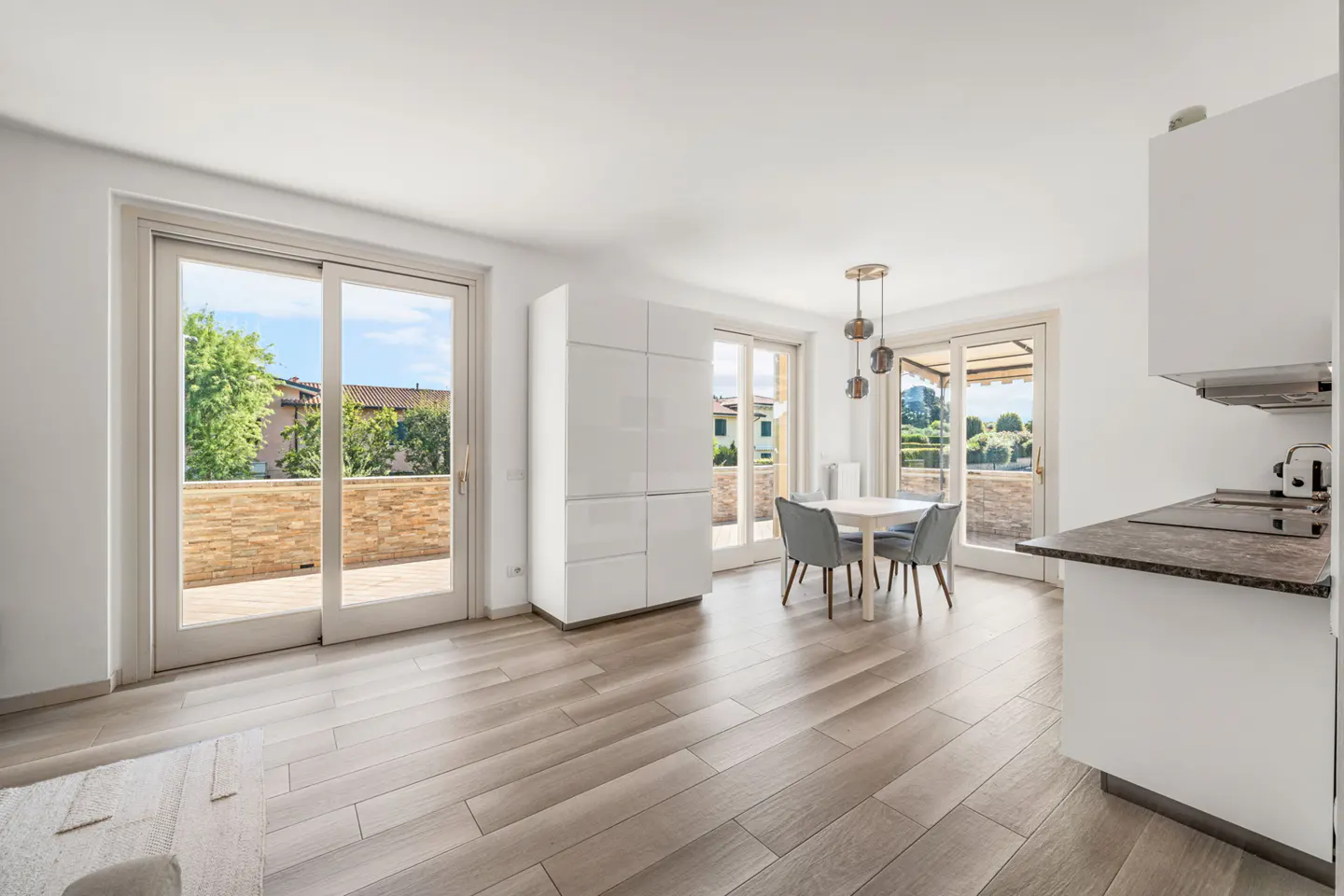 Bright, modern apartment interior with wood-look floors, white walls, and sliding glass doors to a balcony. A dining table and kitchen area are visible.