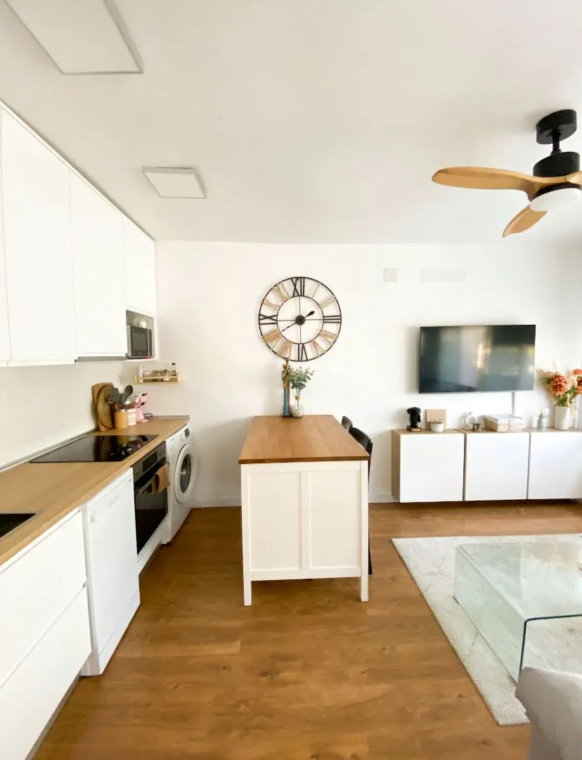 Modern open-plan kitchen and living area with white cabinets, wood-topped island, large wall clock, and wall-mounted TV.