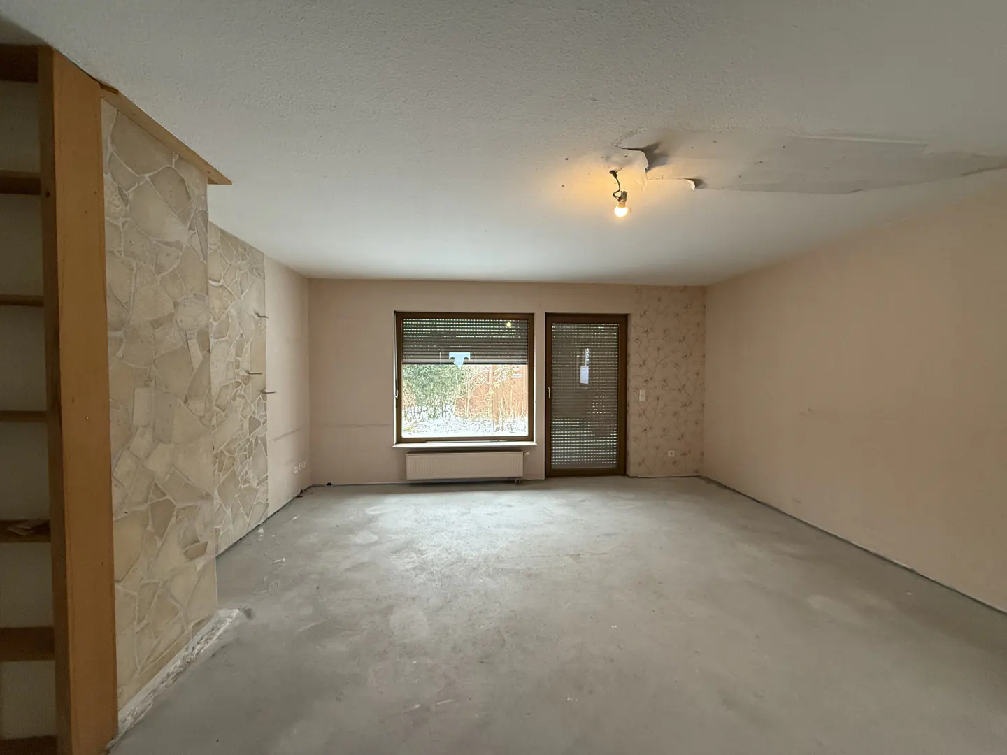 Unfurnished room with beige walls, a stone accent wall, and a window with brown blinds. A single lightbulb hangs from the damaged ceiling.