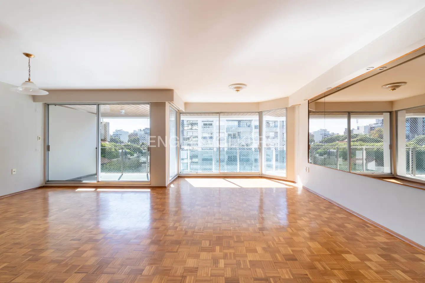 Bright, empty living room with parquet floors, white walls, and large windows overlooking a city and water view. A large mirror reflects the view.