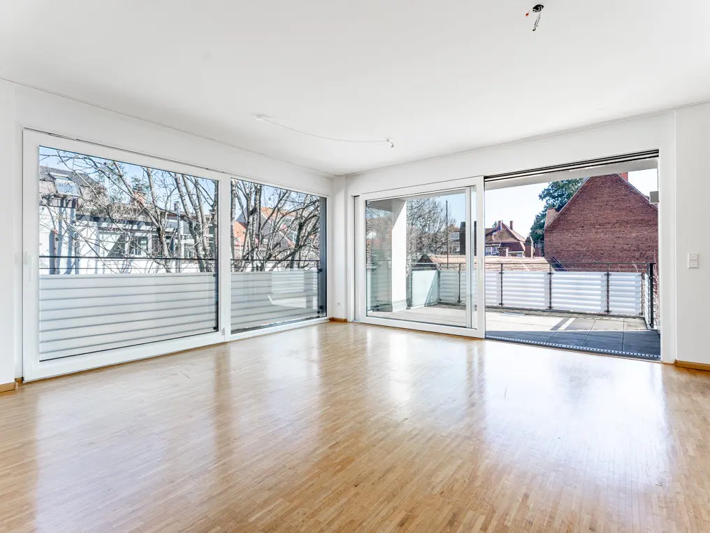 Bright, empty room with light wood floors, white walls, and large windows. Balcony access with sliding glass doors.