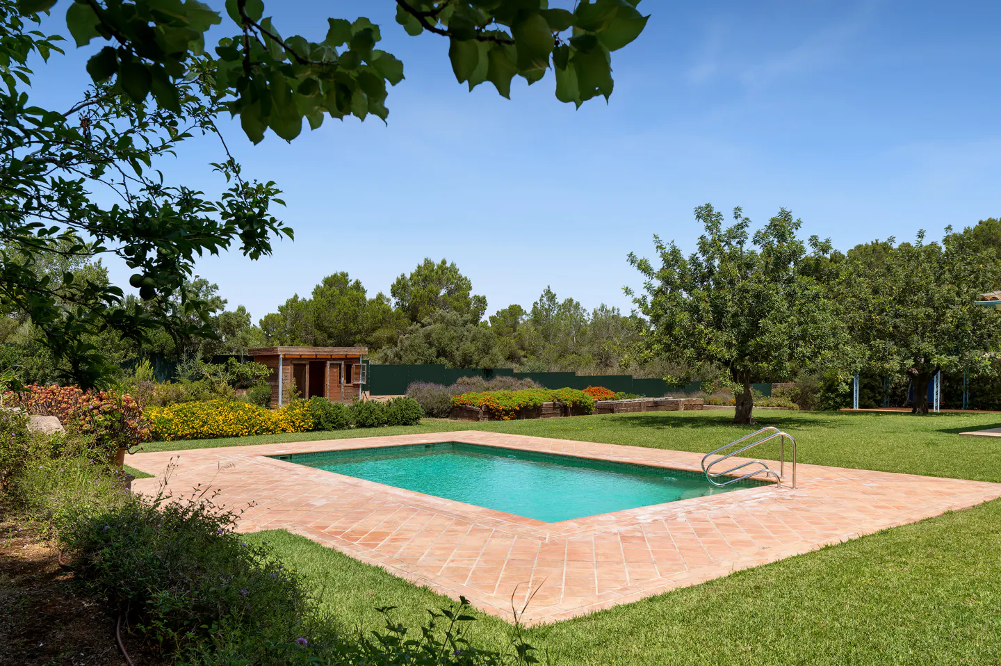 A rectangular swimming pool with turquoise water and a terracotta tile deck, set in a lush green garden under a blue sky.
