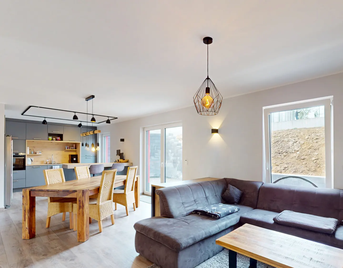 Open-concept living space with a gray sectional sofa, wood dining table with wicker chairs, and a modern gray kitchen.