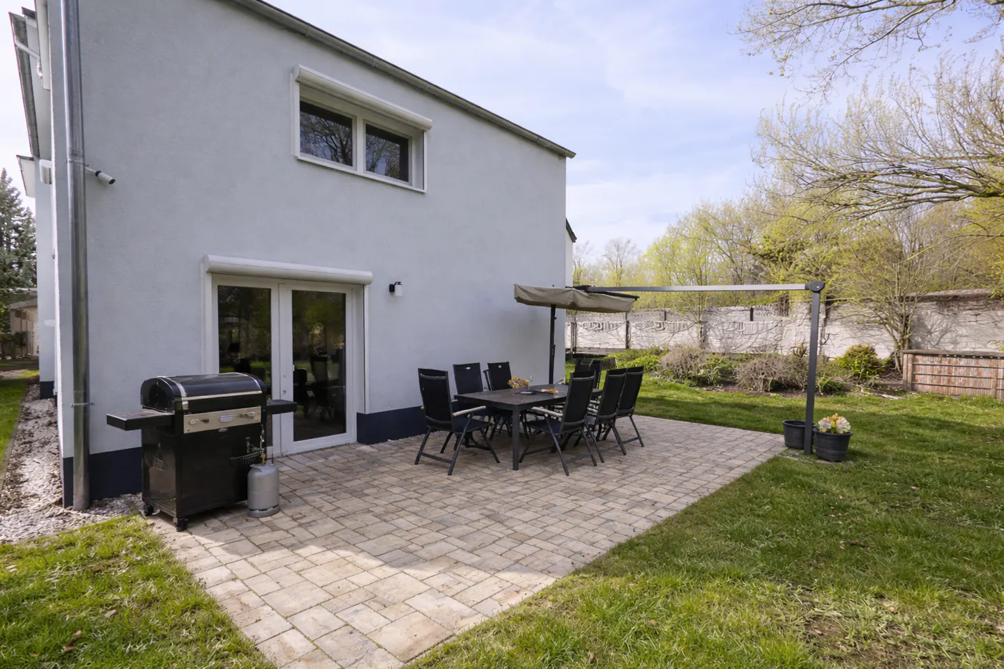 Backyard patio with a grill, table, and chairs. The patio is made of light brown pavers and is surrounded by green grass. A light gray house is in the background.