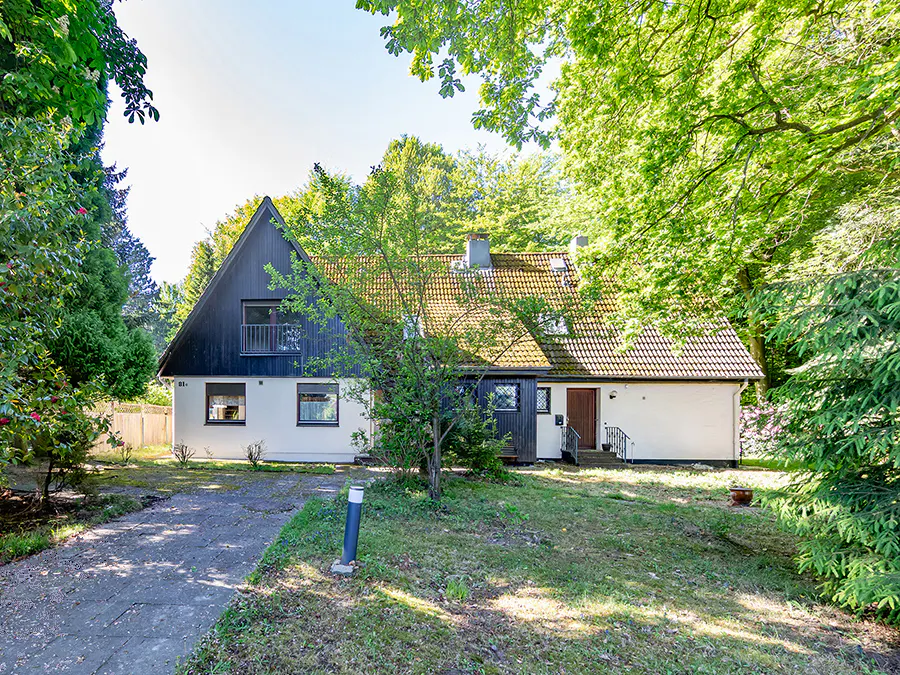 A two-story house with white walls, dark wood accents, and a tiled roof, surrounded by lush green trees and a front lawn.