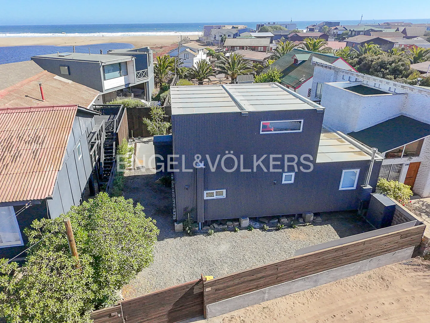 Aerial view of a modern, dark gray house with a flat roof, surrounded by a gravel yard and a wooden fence, near a beach.
