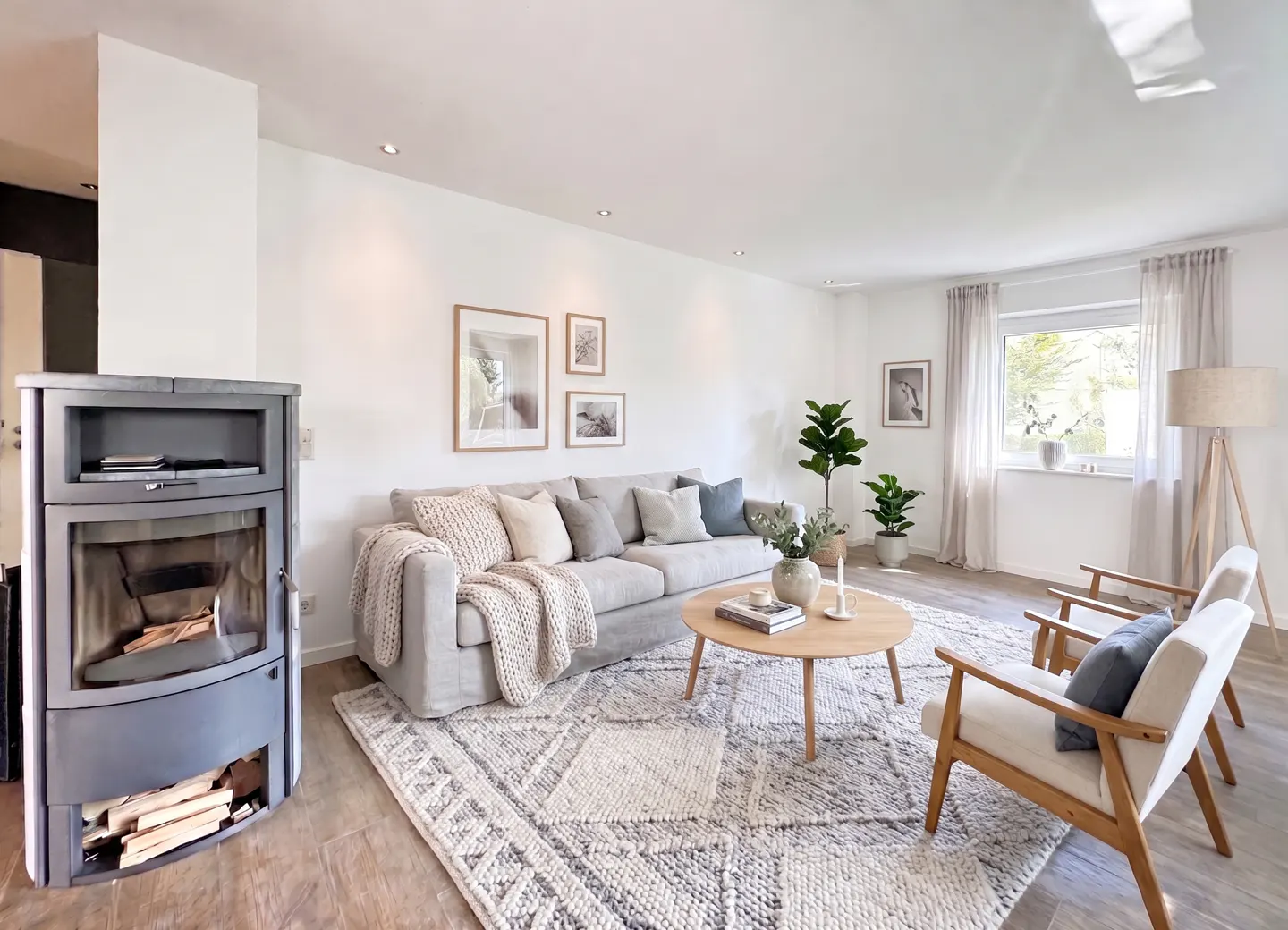 Bright living room with a gray sofa, wood-burning stove, and a round coffee table on a patterned rug. Two chairs face the sofa.
