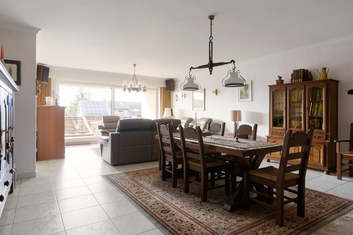 Bright living and dining area with a dark wood table, chairs, and china cabinet on a patterned rug. A brown leather sofa sits near a balcony.