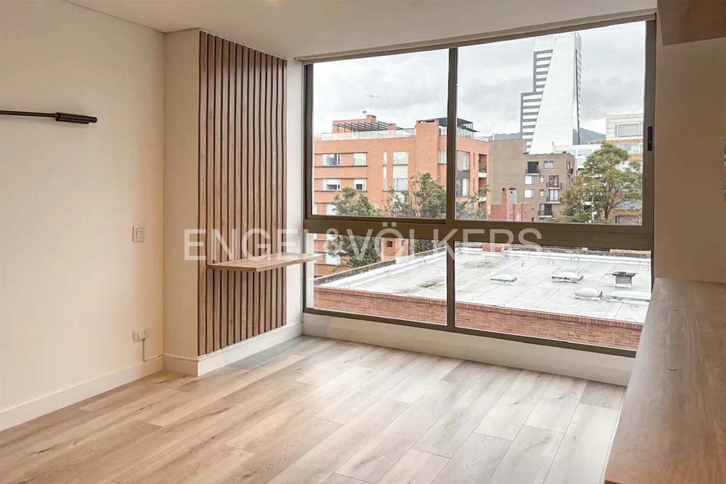 Bright, empty room with wood floors, a large window with a city view, and a small wooden desk attached to a wood-paneled wall.