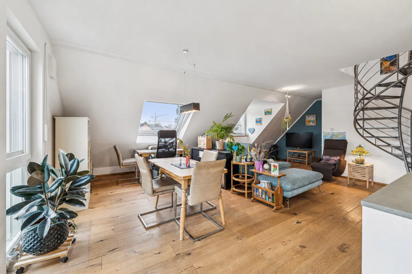 Bright attic living room with wood floors, dining area, blue sofa, TV, and a black metal spiral staircase.