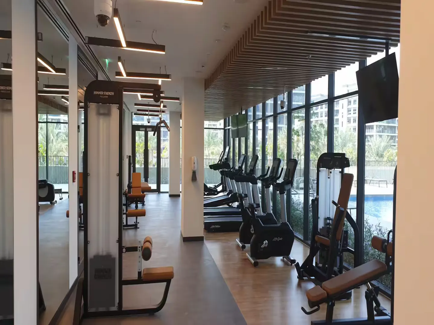 Bright, modern gym with treadmills, bikes, and weight machines. Large windows overlook a pool and city views.