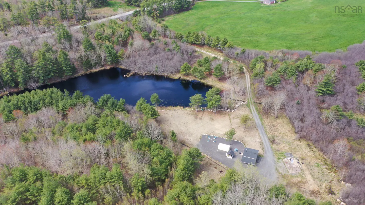 Aerial view of a property with a dark blue pond, surrounded by trees, and a house with a black roof. A dirt road leads to a green field.