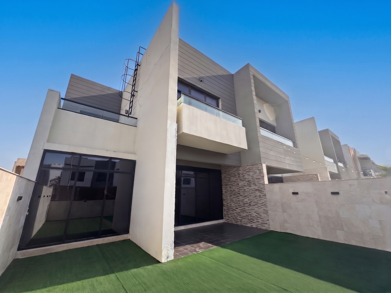 Modern two-story townhouse with a beige facade, glass balconies, large dark windows, and a private green lawn.