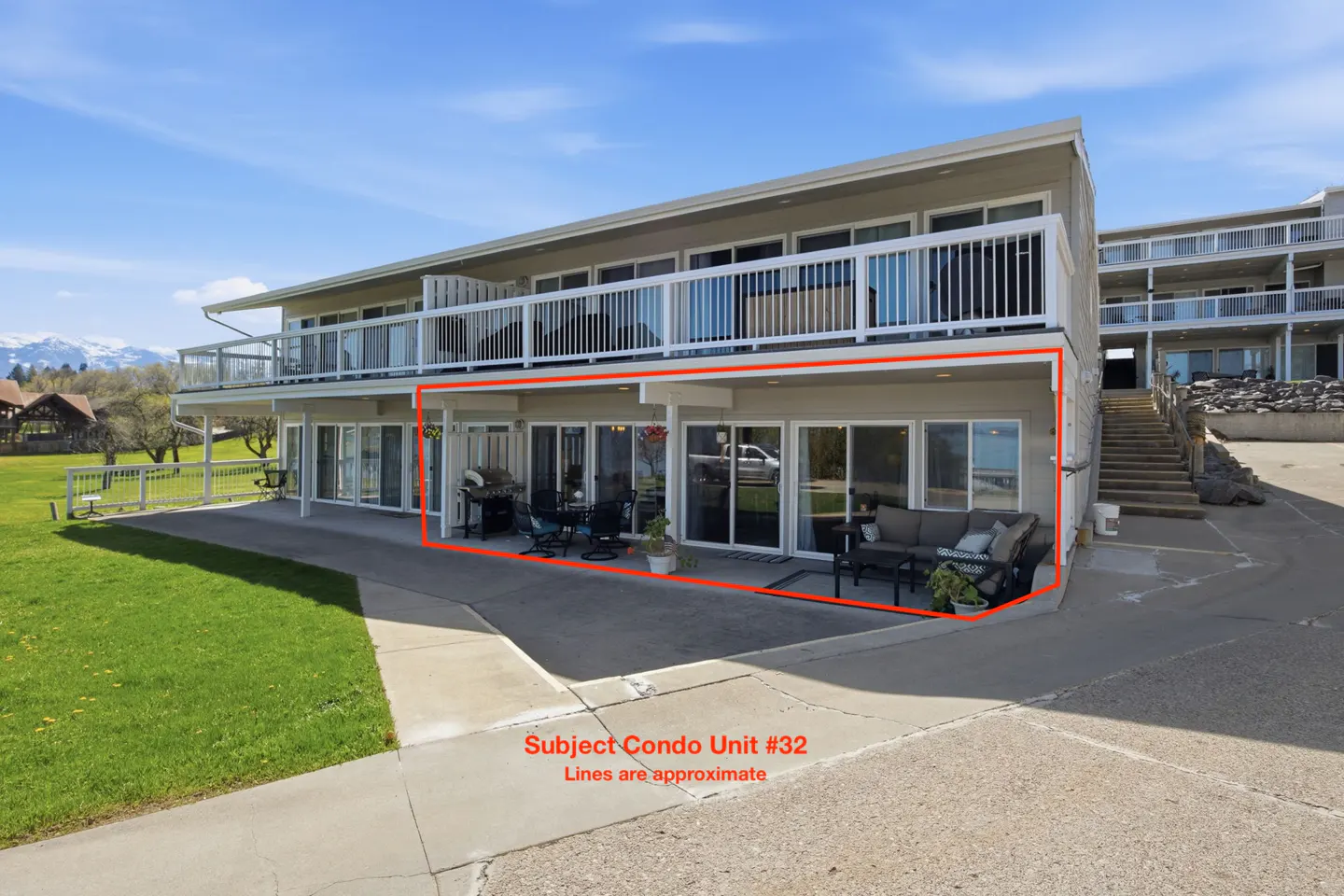 Condo unit #32 with a patio, grill, and outdoor seating. The building is two stories with white railings and large windows. Mountains in the background.