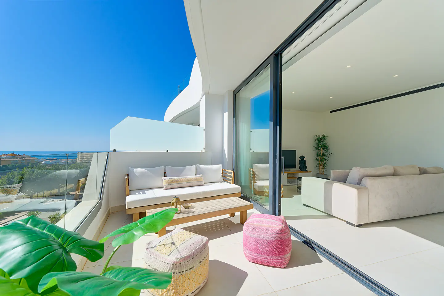 Bright, modern balcony with a daybed, coffee table, and colorful poufs. Sliding glass doors open to a living room with a sofa. Ocean view.