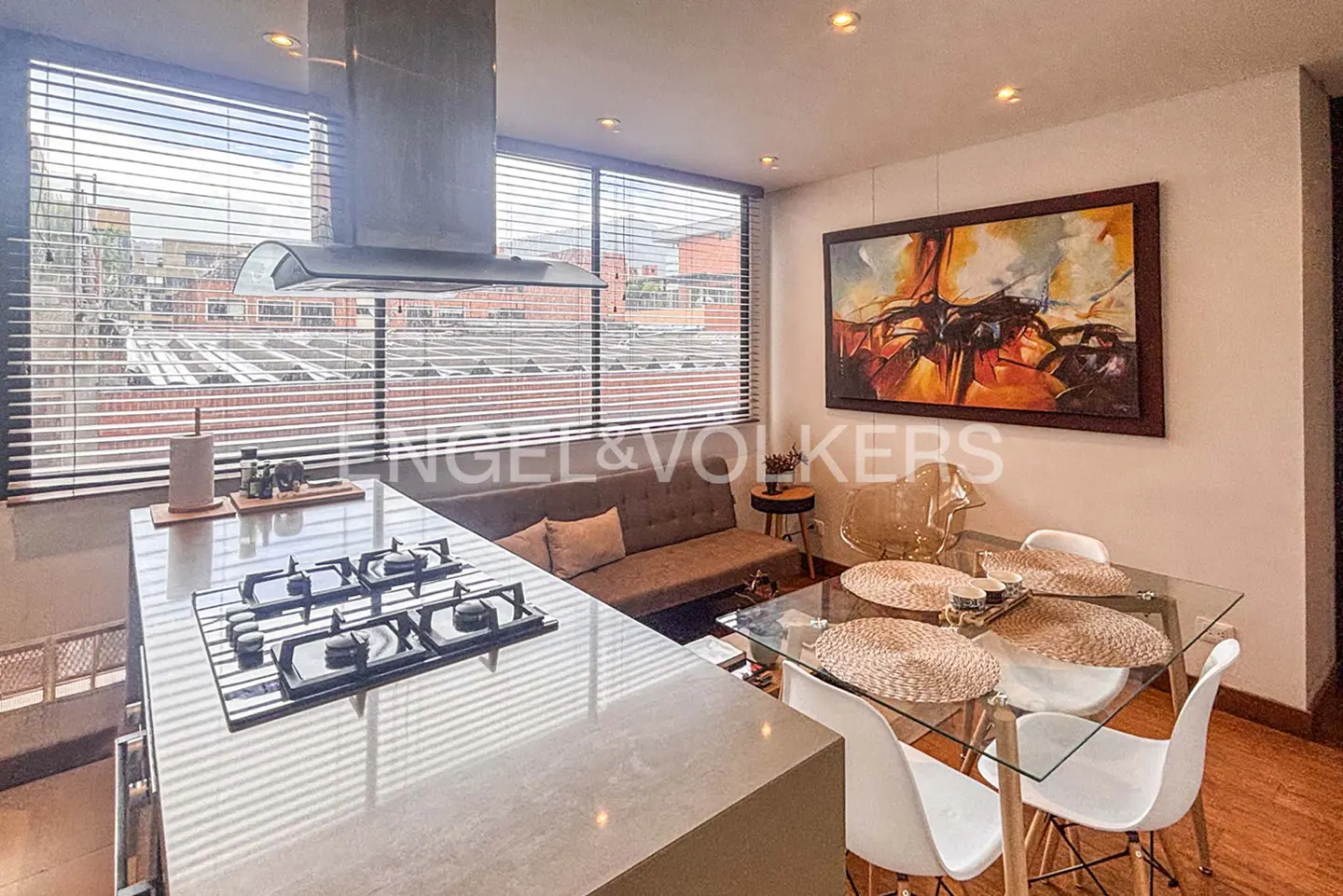 A modern apartment interior with a kitchen island, dining table, and sofa, lit by natural light from large windows.