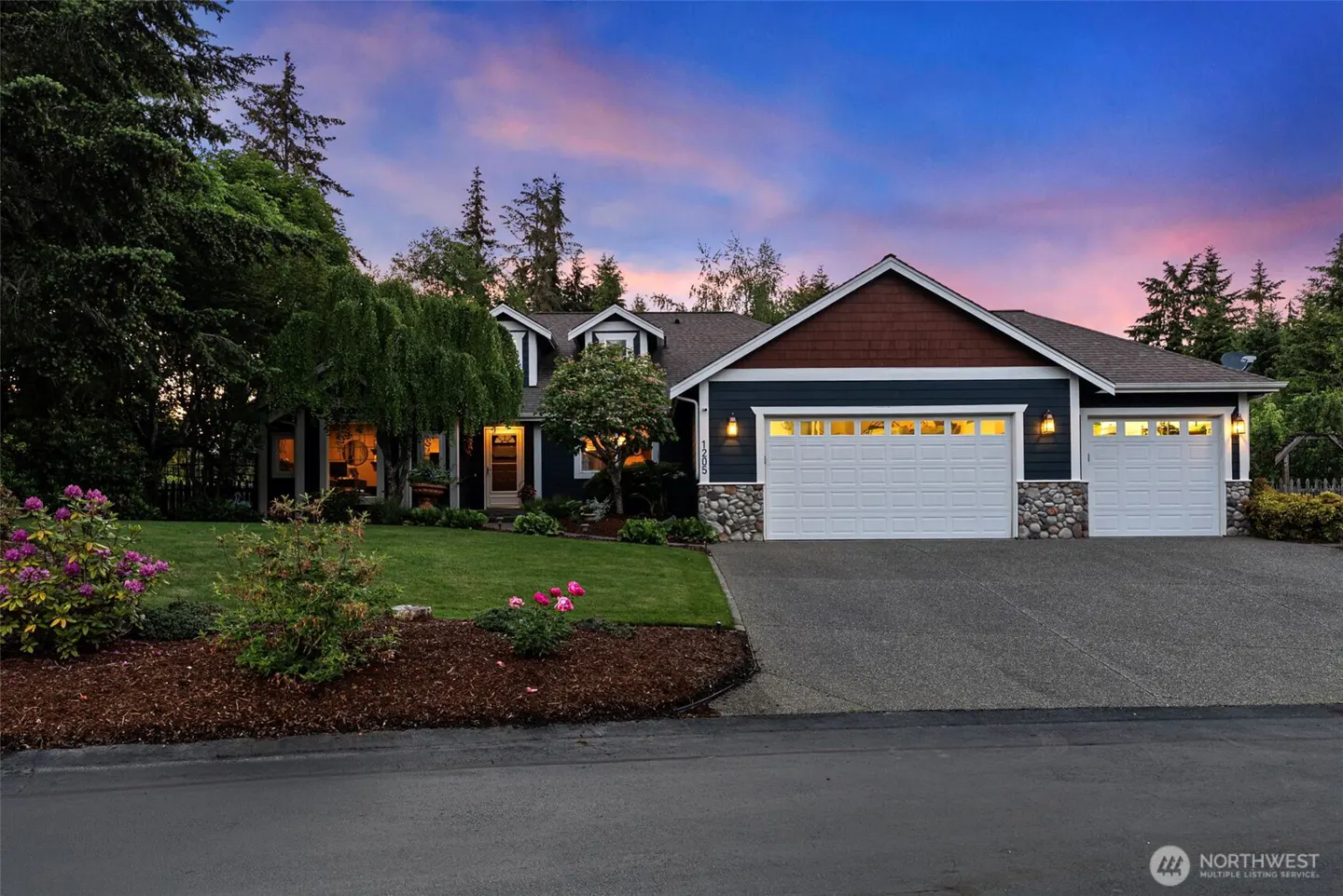 Exterior shot of a two-story house with a two-car garage, blue siding, and a brown roof, surrounded by green trees and a lawn.