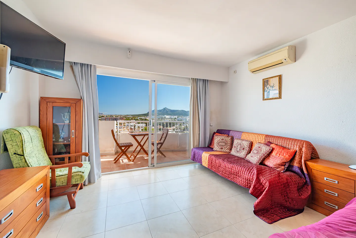 Bright living room with balcony. A sofa with colorful pillows, wooden furniture, and a view of the city and mountains.