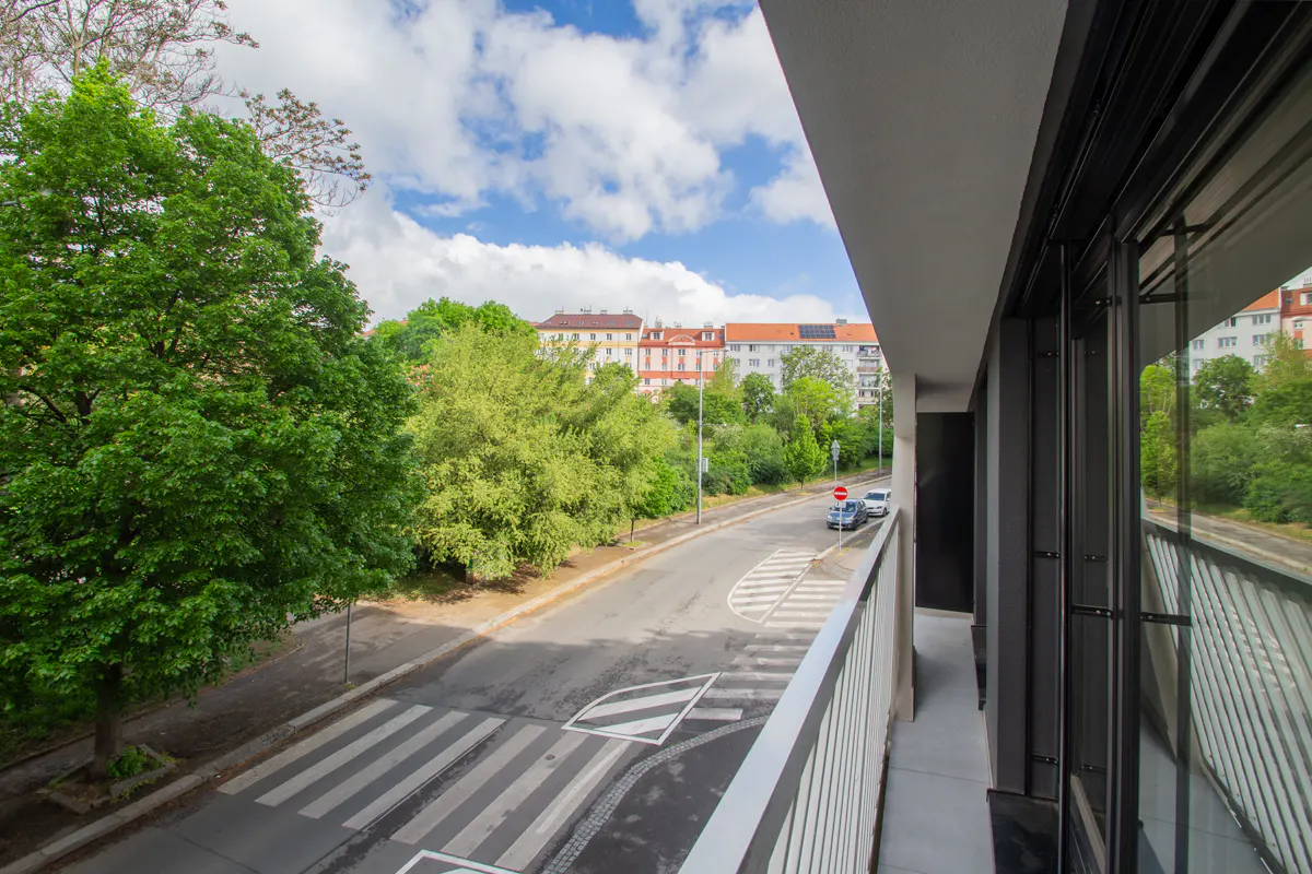 View from a balcony overlooking a street with a crosswalk, trees, and buildings under a blue sky with clouds.