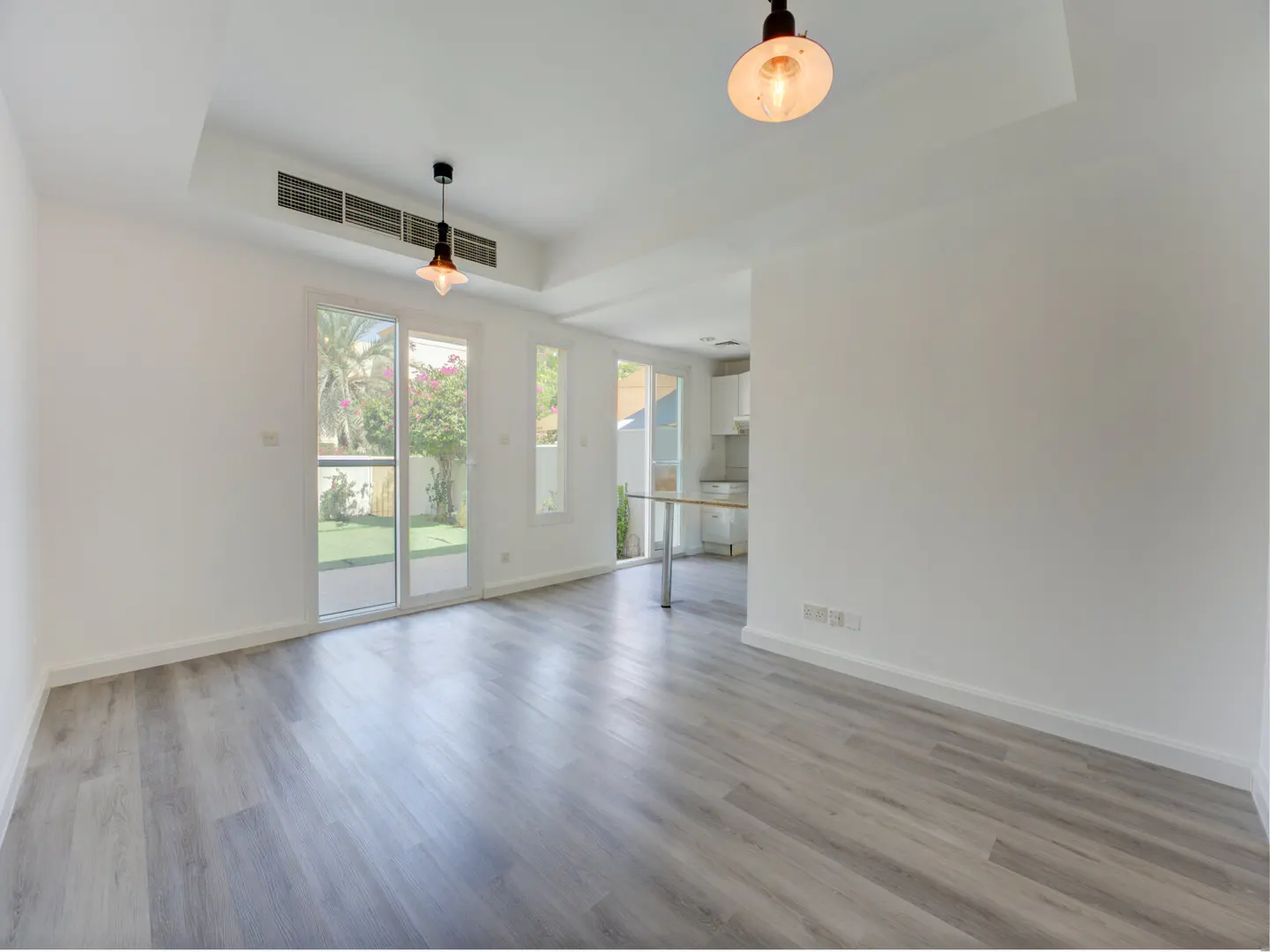 Bright, empty room with gray wood floors, white walls, and two pendant lights. Sliding glass doors lead to a green lawn and garden.