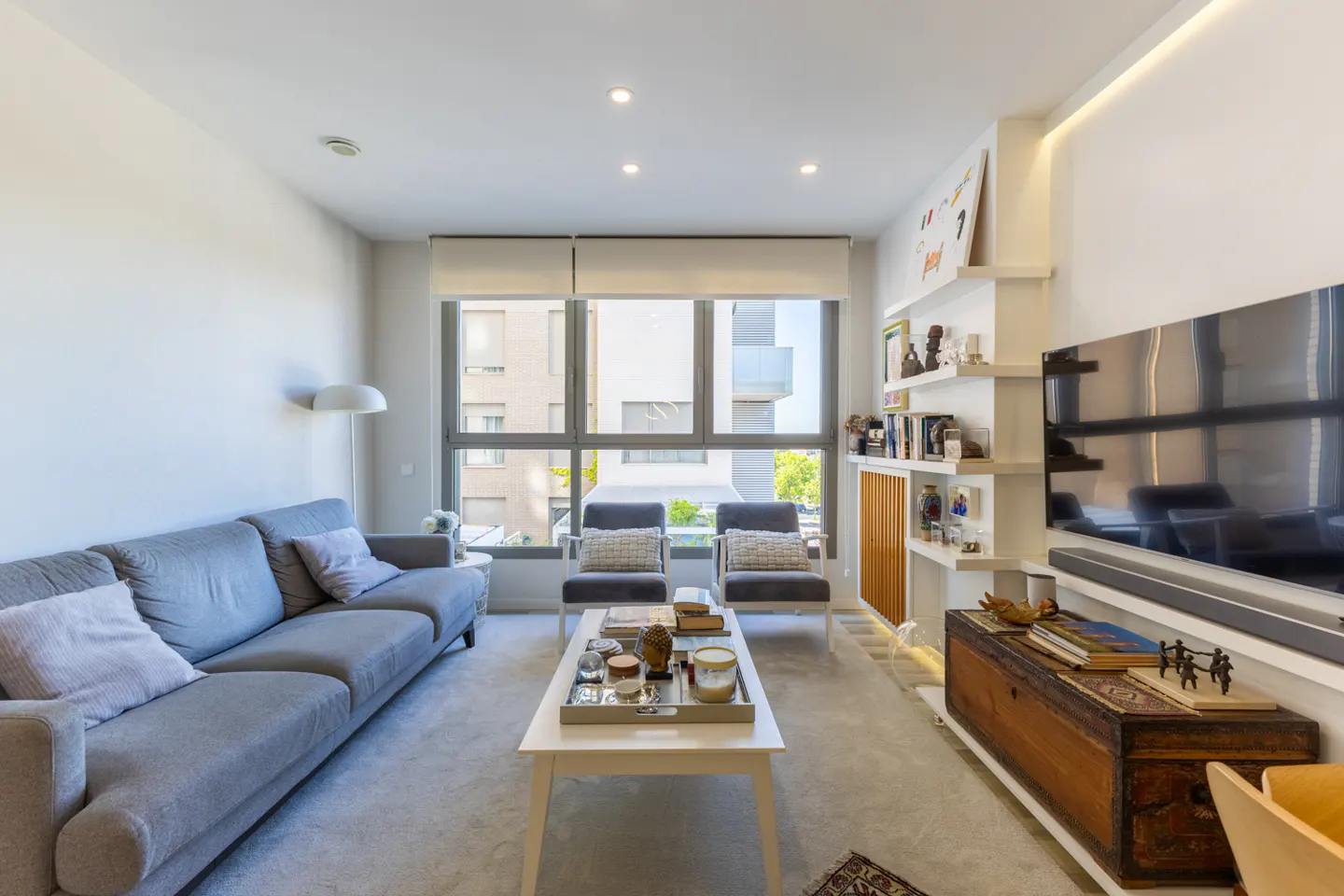 Modern living room with a grey sofa, two armchairs, a white coffee table, and a large wall-mounted TV over a wooden chest.