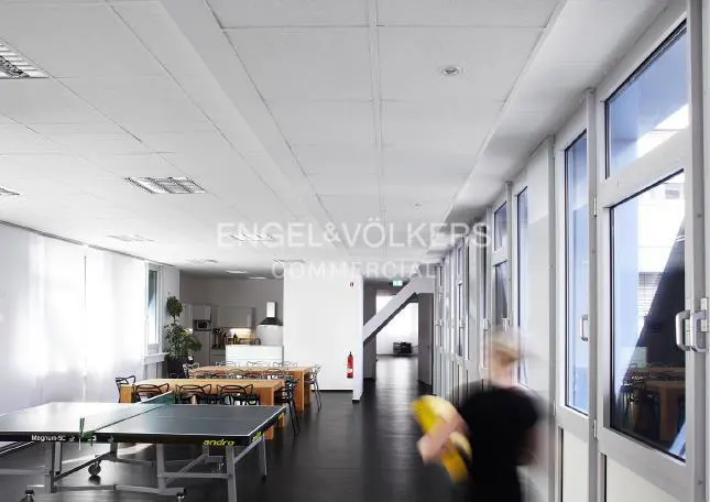 Bright office space with ping pong tables, wooden tables and chairs, and a blurred person holding a yellow object.