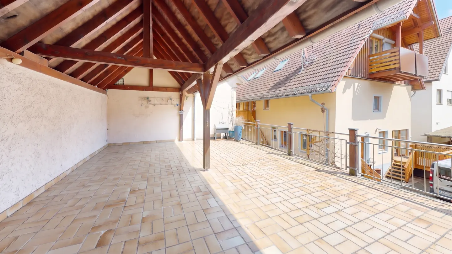 A large, open-air patio with a wooden roof structure and beige brick flooring. A railing separates the patio from the adjacent building.