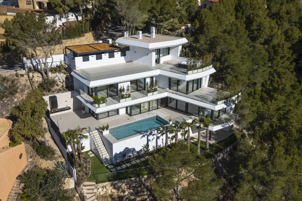 Aerial view of a modern, multi-story white house with a pool, surrounded by trees on a sunny day.
