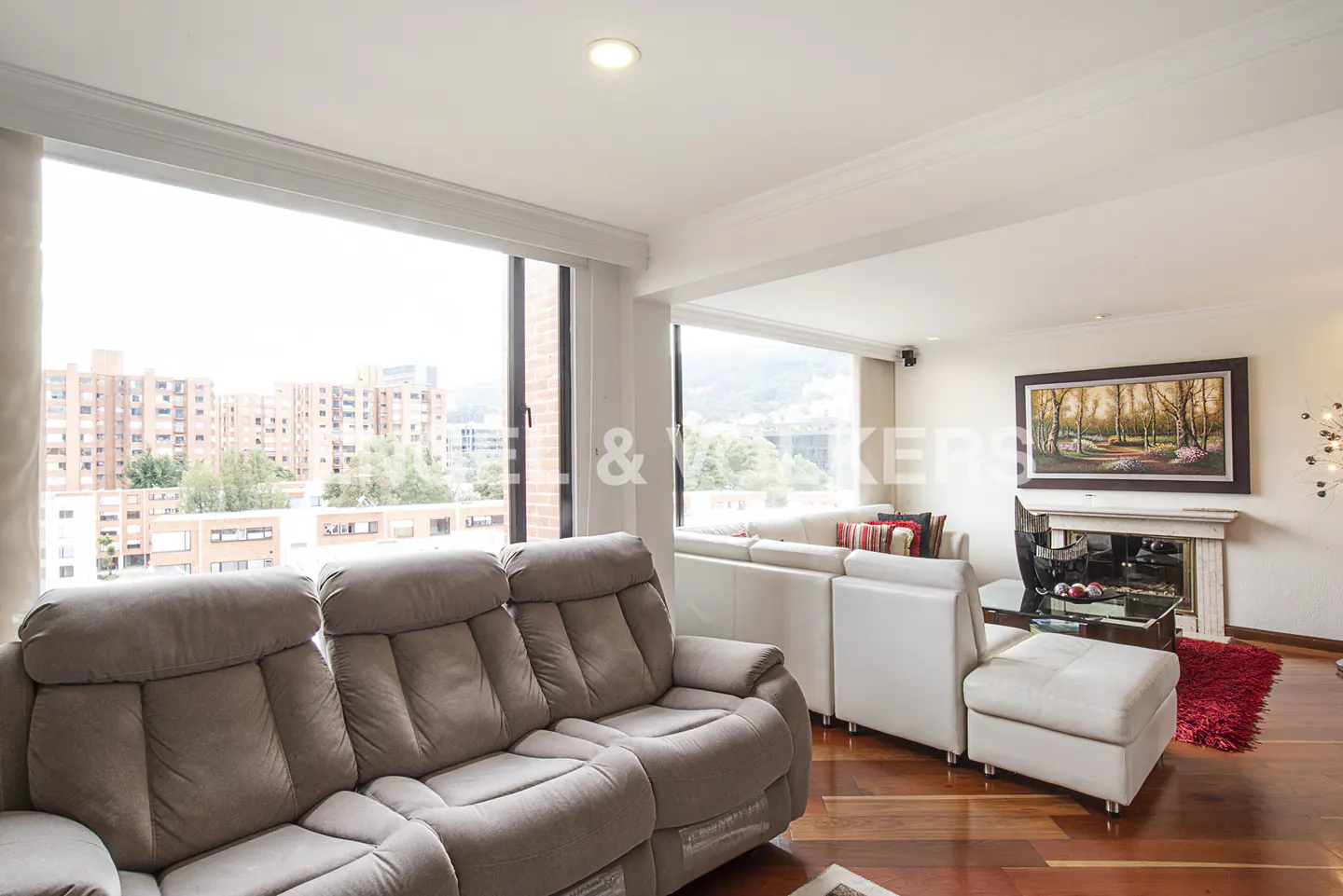 Bright living room with a grey reclining sofa, white sectional, fireplace, and large windows overlooking city buildings.
