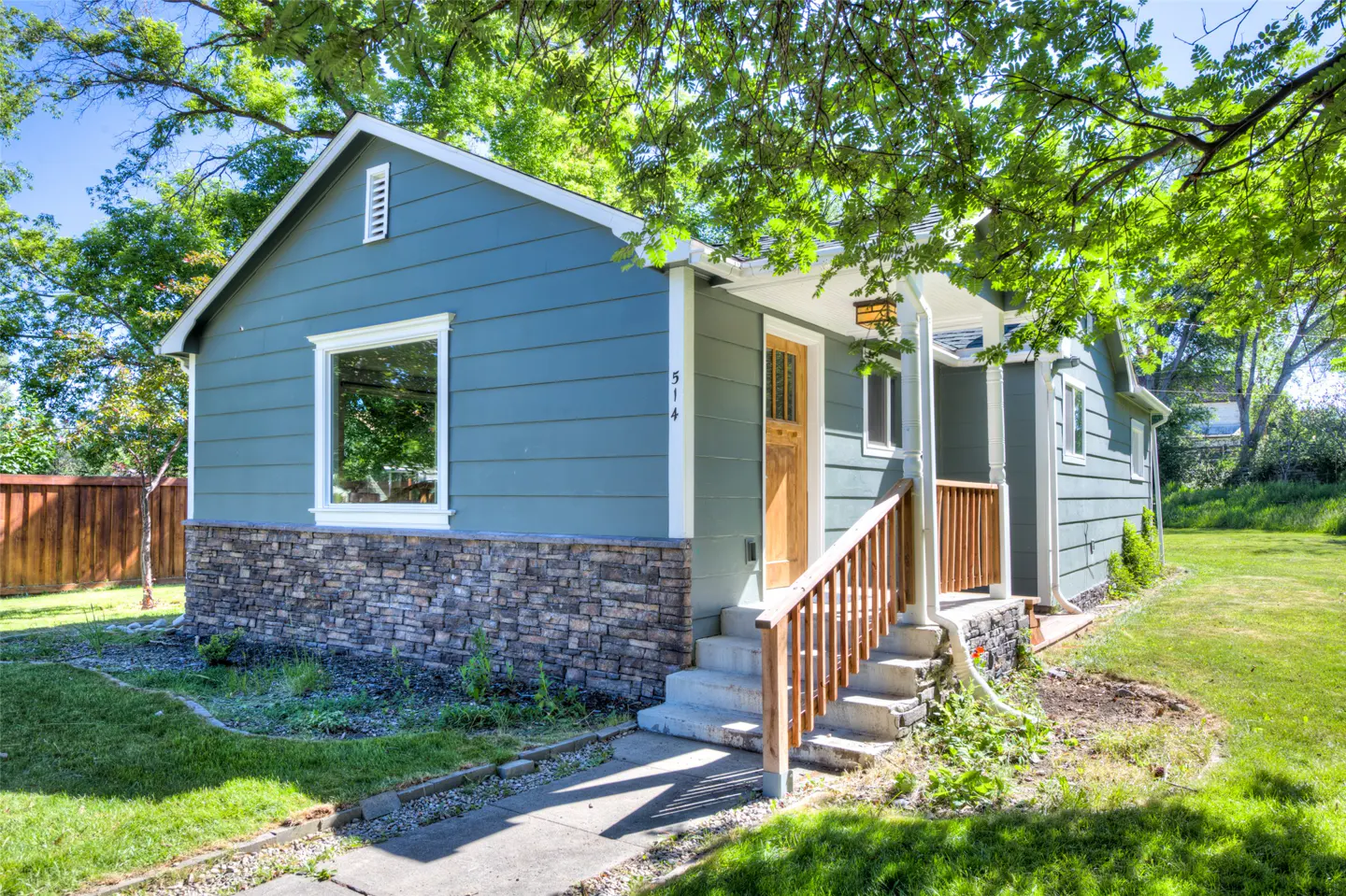 Blue house with stone siding, a wooden front door, and a small porch, surrounded by a green lawn and trees.