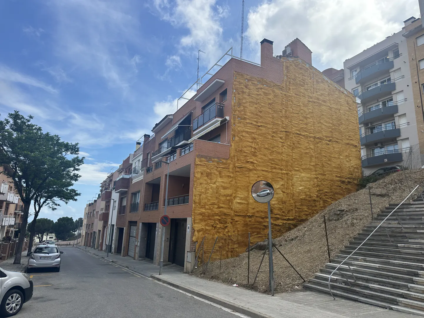 Street view of a multi-story brick apartment building with balconies and a yellow textured wall. Stairs lead up a hill to another building.