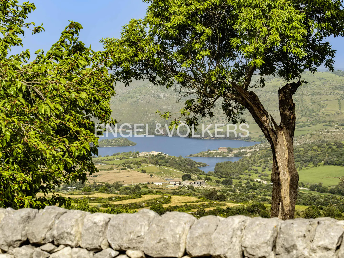 A stone wall in the foreground overlooks a lush green valley with a blue lake and distant hills under a clear blue sky.