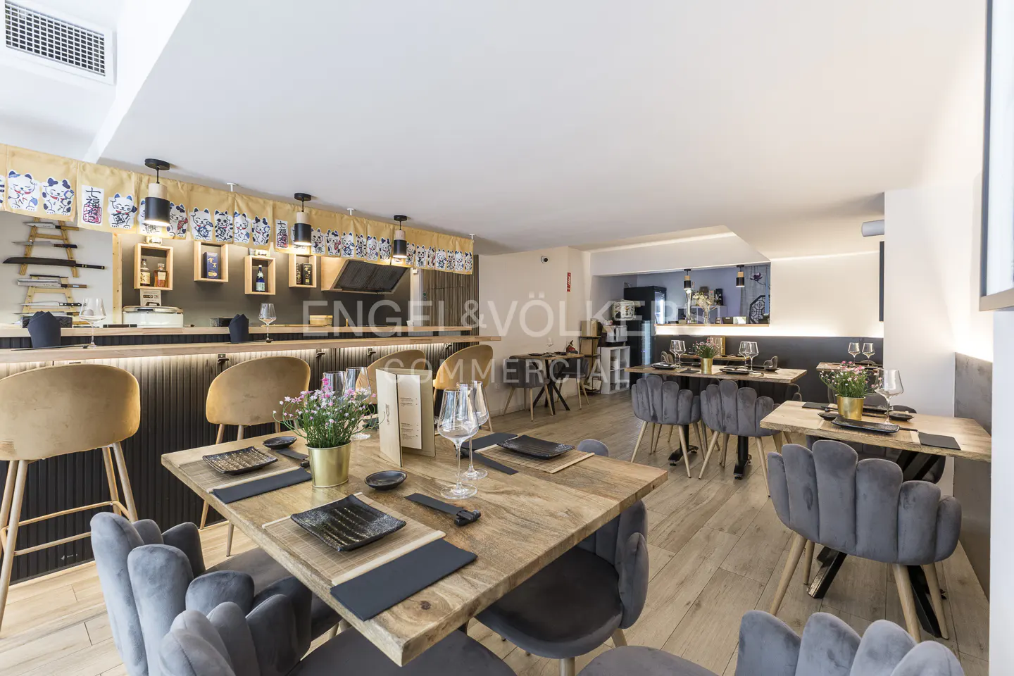 Restaurant interior with wooden tables, gray velvet chairs, and a bar with tan stools. Japanese decor and lighting. Engel & Völkers logo.