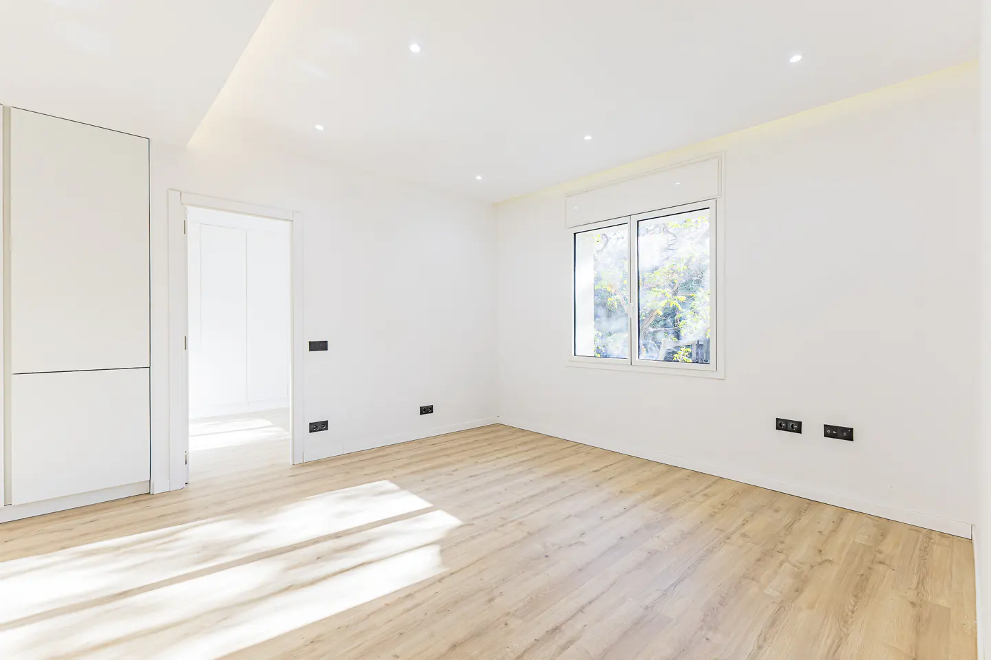 Bright, empty room with light wood floors, white walls, and a window showing trees outside. A white closet and doorway are on the left.