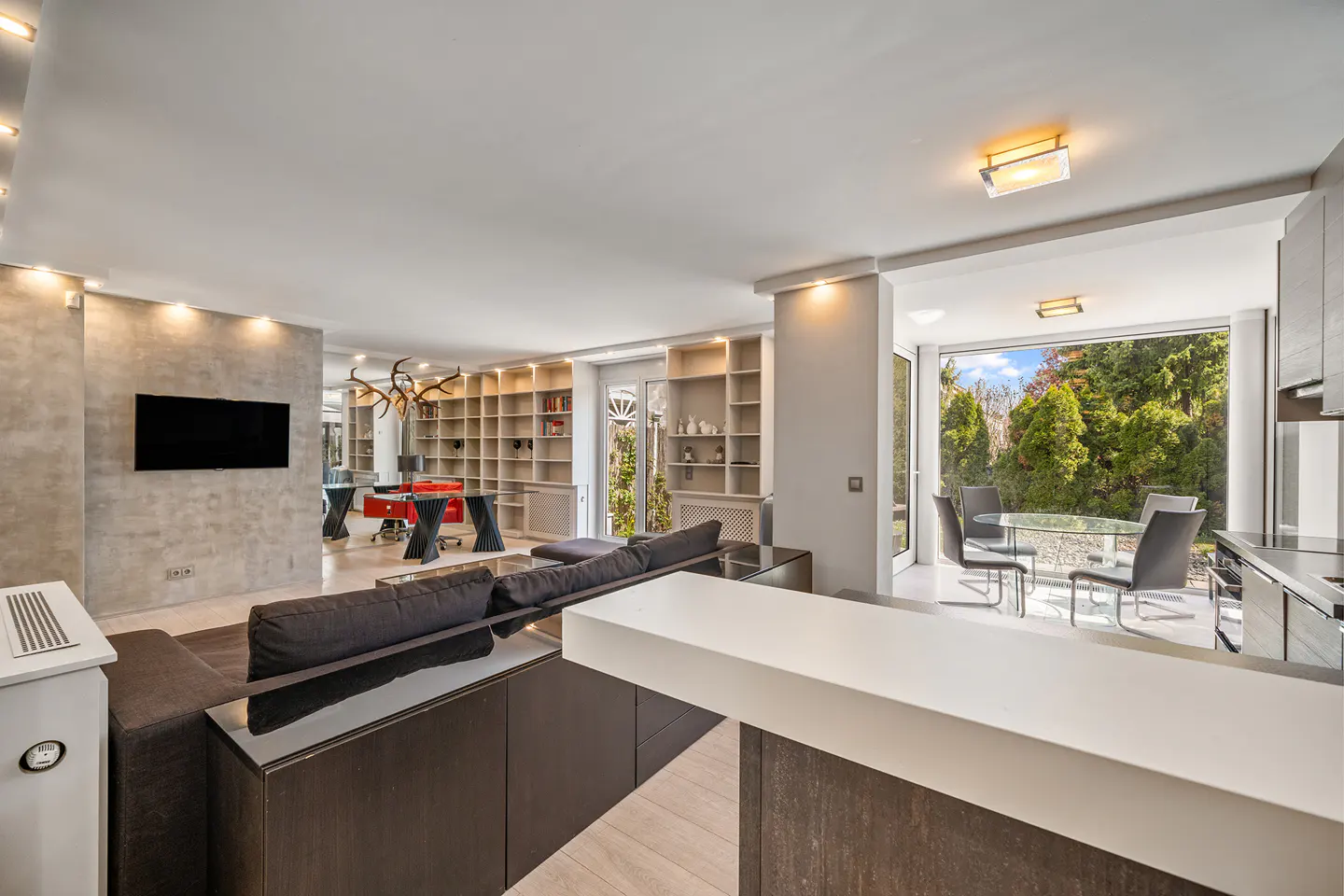 Open-concept living space with a dark sofa, white bar, and a dining area with a view of lush greenery.