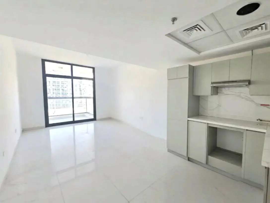 Bright, empty apartment with white walls, tile floors, and a black-framed window. Gray kitchen cabinets line the right wall.