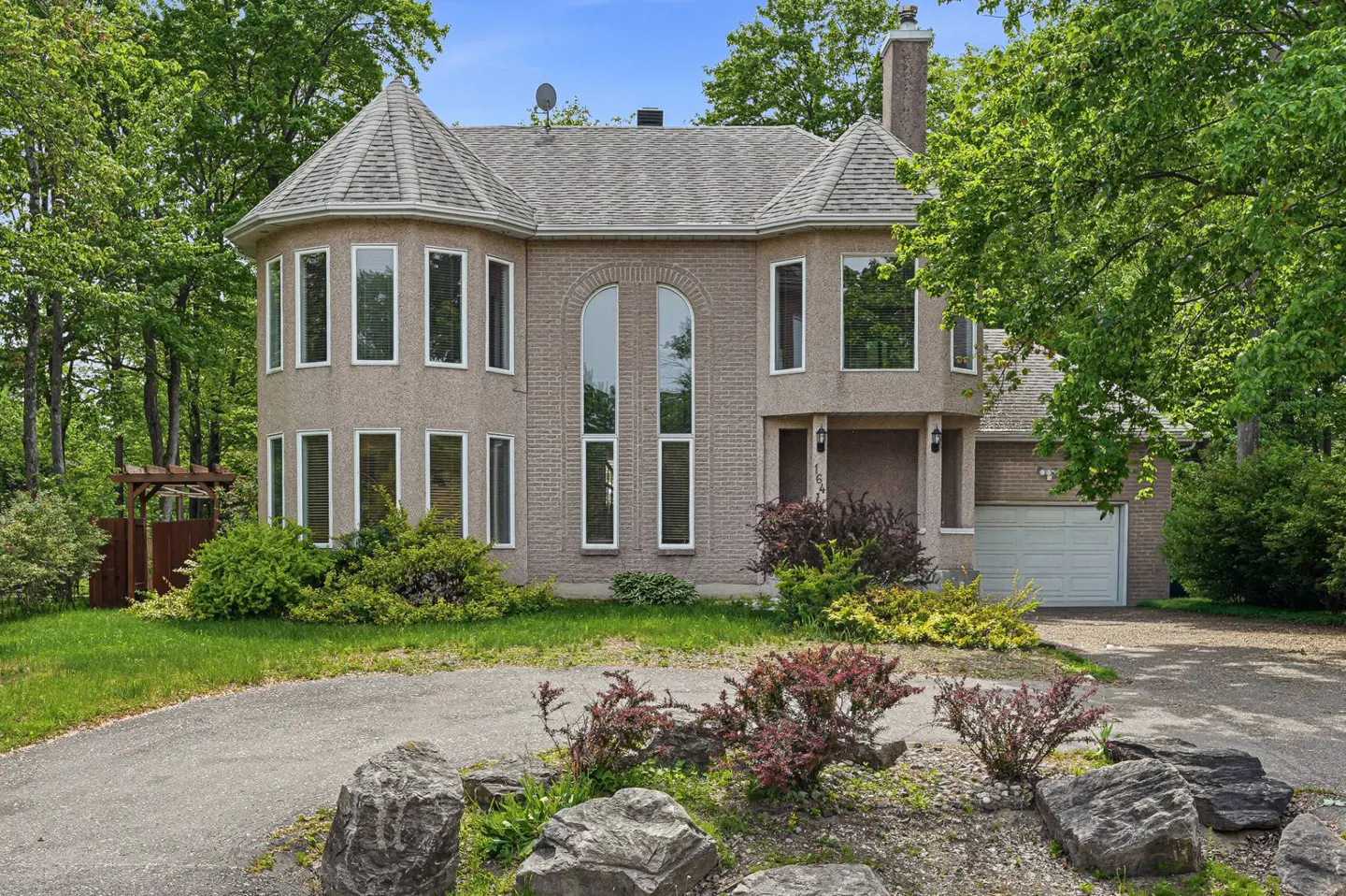 Two-story tan brick house with turrets, arched windows, and an attached garage, surrounded by lush trees and landscaping.