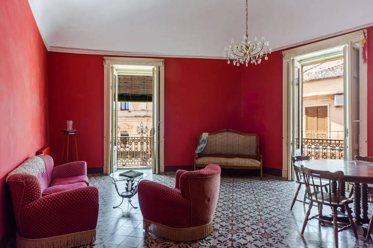A red living room with a sofa, chair, table, and chandelier. Balconies are visible through open doors.