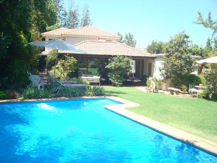 Backyard view of a two-story house with a pool, lawn, patio, and trees. The pool is turquoise. The house has a tan tile roof.