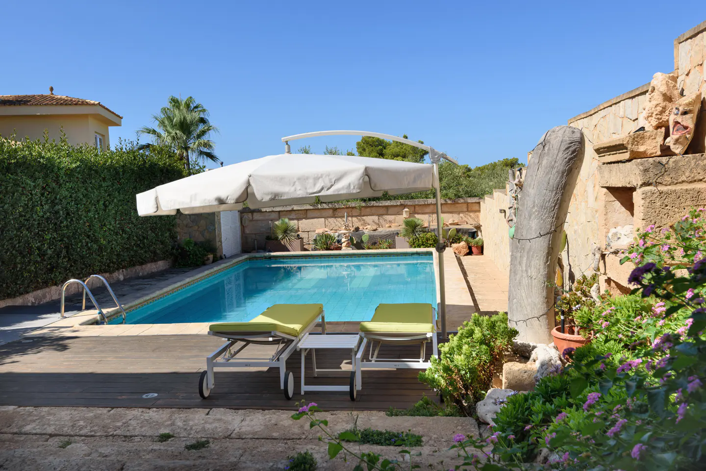 A backyard pool with two lounge chairs under a white umbrella, surrounded by greenery and a stone wall.